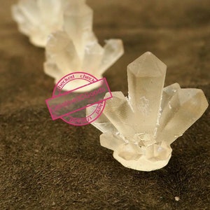 May include: Three clear crystal clusters of varying sizes, arranged in a row on a brown textured surface.