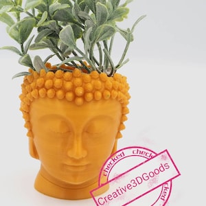 Puede incluir: Un pequeño macetero de cabeza de Buda naranja con una planta verde frondosa en su interior. El macetero tiene una superficie texturizada con pequeños bultos. La cabeza de Buda tiene los ojos cerrados y una expresión serena.