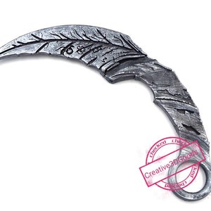 Könnte beinhalten: Ein silberfarbenes Wurfmesser aus Metall mit einer gebogenen Klinge und einem detaillierten Design. Die Klinge hat eine strukturierte Oberfläche und einen Ring am Ende des Griffs.