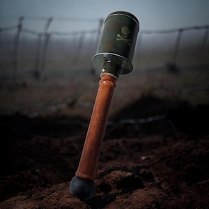 Könnte beinhalten: Eine Handgranate im Vintage-Stil mit Holzgriff und dunkelgrünem Metallaufsatz. Auf dem Aufsatz befindet sich ein Totenkopf- und gekreuzte Knochen-Symbol. Die Granate ist vor einem verschwommenen Hintergrund aus Stacheldraht und Erde platziert.