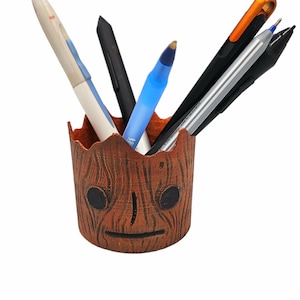 May include: A brown wooden pencil holder shaped like the character Groot from the Guardians of the Galaxy movie. The holder has a black painted face with a serious expression. Five pens are sticking out of the top of the holder, including a blue pen, a black pen, a silver pen, and two orange pens.