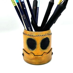 May include: A yellow and brown ceramic pencil holder shaped like a face with black eyes. The holder is filled with various art supplies, including paintbrushes, pencils, and a stylus.
