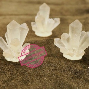 May include: Three clear crystal clusters, each with a different shape and size, are arranged on a brown surface. The crystals are made of clear resin and have a smooth, polished finish.
