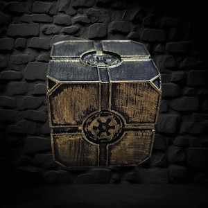 May include: A metallic, box-shaped object with a weathered gold and black finish. The box features a geometric design with a circular emblem on the front. The object is set against a dark, textured background.