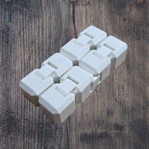 Pode incluir: Um brinquedo fidget branco articulado, composto por vários segmentos cúbicos conectados. O brinquedo foi projetado para dobrar e flexionar, oferecendo uma experiência tátil. O fundo é uma superfície de madeira.