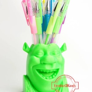 Puede incluir: Un porta lápices verde con temática de Shrek con una amplia sonrisa y una hilera de dientes. El porta lápices está lleno de bolígrafos de colores.