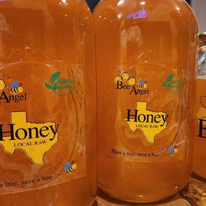 以下が含まれることがあります： 金色の蜂蜜が入ったガラス瓶。各瓶には「Bee Angel Honey」と「Local Raw」の文字とテキサスの輪郭が描かれたラベルが付いています。ラベルには「Plant a tree, save a bee」の文字も含まれています。
