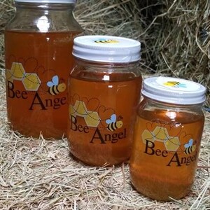 May include: Three glass jars of honey with white lids, varying in size, are displayed. Each jar has a label with the text "Bee Angel" and a bee graphic. The honey is a golden amber color, and the jars are set against a background of hay.