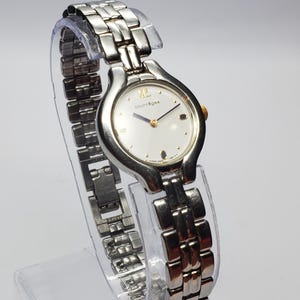 Puede incluir: Un reloj de pulsera Courr&egrave;ges plateado con esfera blanca y detalles dorados. El reloj presenta una correa de metal con eslabones y una esfera redonda con el nombre de la marca. El reloj se exhibe sobre un soporte transparente.