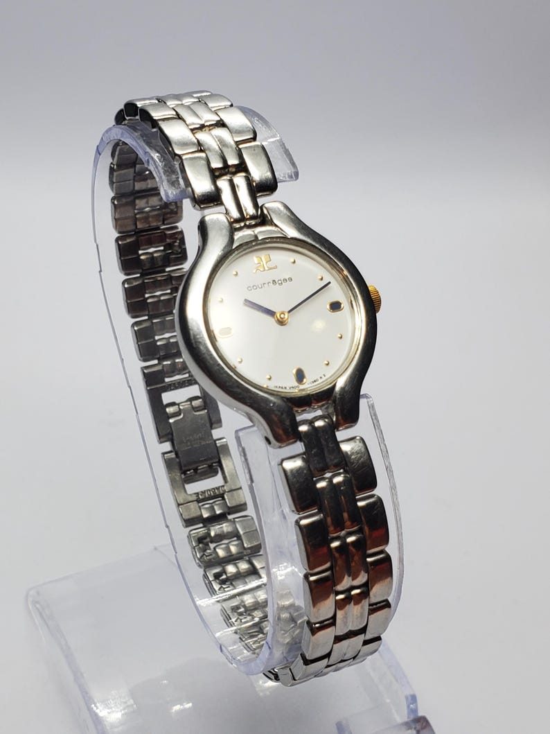 Puede incluir: Un reloj de pulsera Courr&egrave;ges plateado con esfera blanca y detalles dorados. El reloj presenta una correa de metal con eslabones y una esfera redonda con el nombre de la marca. El reloj se exhibe sobre un soporte transparente.