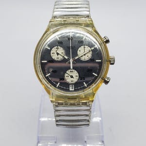 May include: A clear and yellow Swatch chronograph watch with a black dial. The watch features white sub-dials, a date display, and a clear plastic band. The watch is displayed on a clear stand.