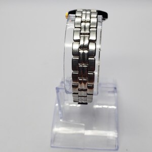 Puede incluir: Un reloj de pulsera plateado con una correa de eslabones rectangulares. La esfera del reloj es negra con detalles dorados. El reloj se exhibe sobre un soporte de pl&aacute;stico transparente sobre un fondo blanco. La correa mide aproximadamente 1,9 cm de ancho.