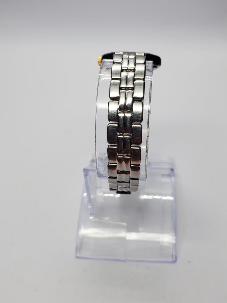Puede incluir: Un reloj de pulsera plateado con una correa de eslabones rectangulares. La esfera del reloj es negra con detalles dorados. El reloj se exhibe sobre un soporte de pl&aacute;stico transparente sobre un fondo blanco. La correa mide aproximadamente 1,9 cm de ancho.