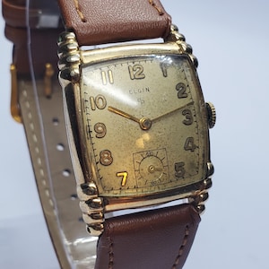 Op de afbeelding: Een vintage Elgin-horloge met een rechthoekige goudkleurige wijzerplaat en een bruine leren band. De wijzerplaat toont het Elgin-logo en cijfers. Het horloge heeft een kleine secondenwijzerplaat en een bruine leren band.