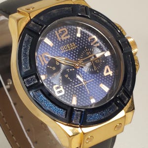 May include: A gold-coloured Guess watch with a black bezel and a blue dial. The watch features gold numerals and hands, and a black leather strap. The watch face displays the word "GUESS" and other details.