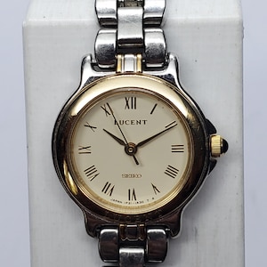 May include: A Seiko Lucent wristwatch featuring a round cream-colored dial with black Roman numeral hour markers. The watch has a two-tone silver and gold-tone metal link bracelet and a gold-tone bezel. The dial displays the text Lucent and Seiko.