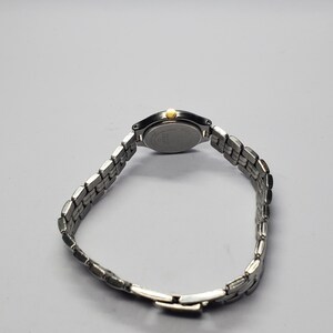 Puede incluir: Un reloj de pulsera plateado con una correa de metal con eslabones. La esfera del reloj es ovalada con una corona dorada. El reloj se muestra sobre un fondo gris claro.