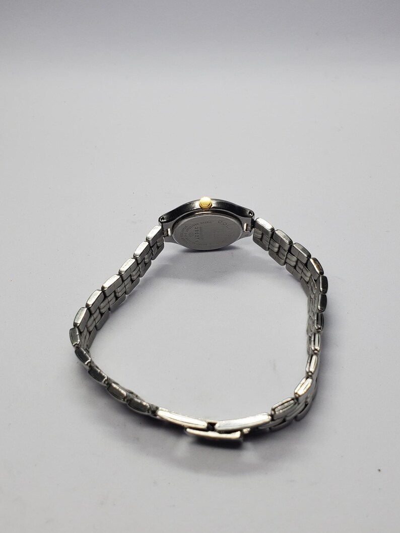 Puede incluir: Un reloj de pulsera plateado con una correa de metal con eslabones. La esfera del reloj es ovalada con una corona dorada. El reloj se muestra sobre un fondo gris claro.