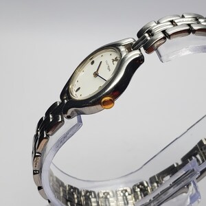 Puede incluir: Reloj de pulsera plateado con esfera ovalada blanca y correa plateada. El reloj tiene una corona dorada y agujas azules. La esfera del reloj tiene el nombre de la marca "courreges".