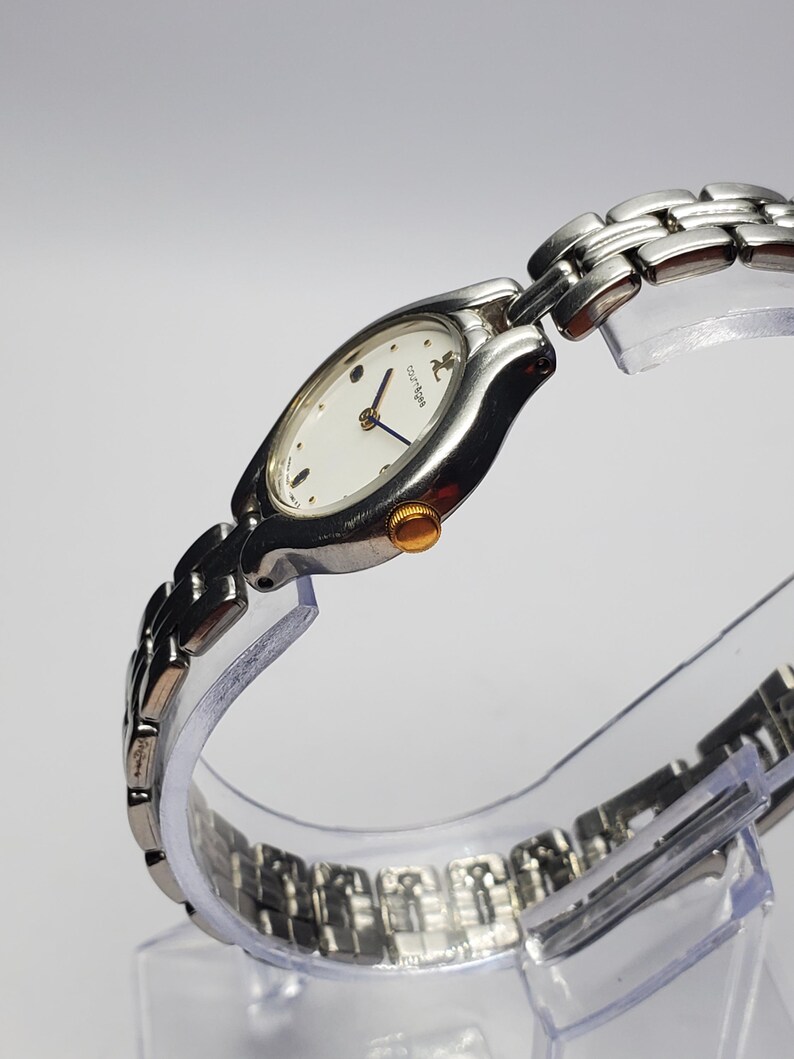 Puede incluir: Reloj de pulsera plateado con esfera ovalada blanca y correa plateada. El reloj tiene una corona dorada y agujas azules. La esfera del reloj tiene el nombre de la marca "courreges".