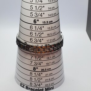 Puede incluir: Una pulsera plateada y marr&oacute;n se coloca en una herramienta de medici&oacute;n EZ Bracelet Mini. La herramienta mide de 12,7 cm a 22,2 cm. La pulsera est&aacute; hecha de eslabones rectangulares.