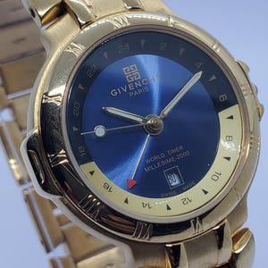 Puede incluir: Reloj Givenchy World Timer Millesime-2000 dorado con esfera azul. El reloj tiene una visualización de la fecha a las 3 en punto y un bisel giratorio con marcas de hora mundial. La correa del reloj también es dorada.
