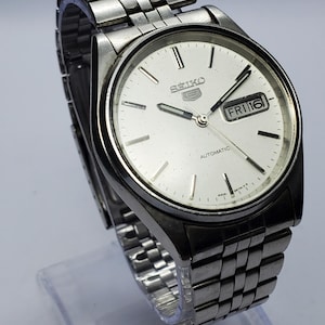 May include: A silver-tone Seiko 5 automatic wristwatch with a round white dial, silver baton hour markers, and a stainless steel link bracelet. The dial features the Seiko logo, the number 5 emblem, and a day-date window displaying FRI 16.