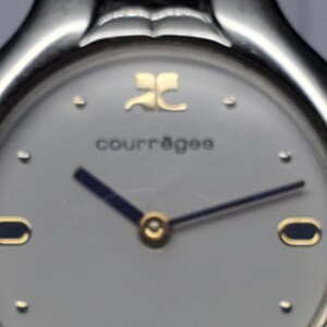 Puede incluir: Primer plano de un reloj de pulsera Courr&egrave;ges. El reloj tiene una caja de metal plateado y una esfera gris claro. Las manecillas son de color azul oscuro con detalles dorados. El nombre de la marca y el logotipo son dorados. Dise&ntilde;o cl&aacute;sico y minimalista.