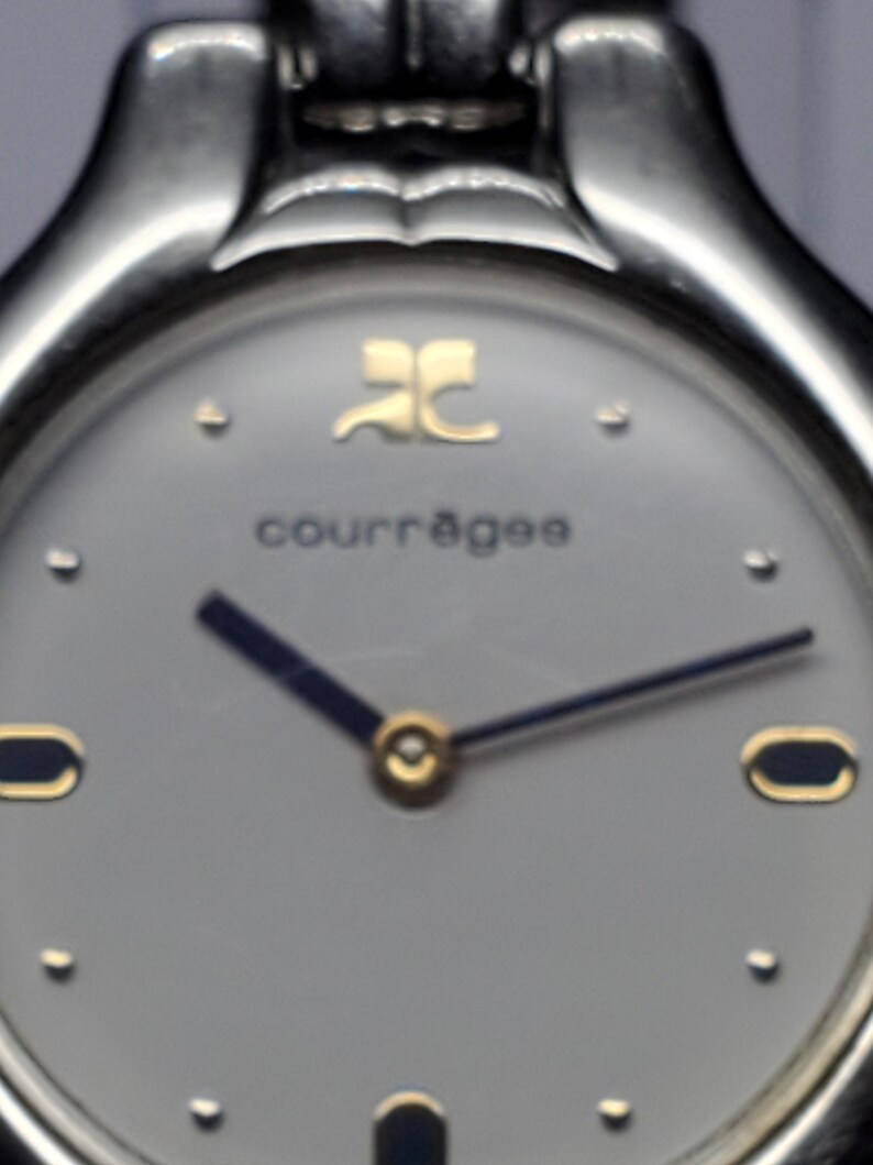 Puede incluir: Primer plano de un reloj de pulsera Courr&egrave;ges. El reloj tiene una caja de metal plateado y una esfera gris claro. Las manecillas son de color azul oscuro con detalles dorados. El nombre de la marca y el logotipo son dorados. Dise&ntilde;o cl&aacute;sico y minimalista.