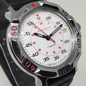 Puede incluir: Un reloj de pulsera plateado con esfera blanca, correa de cuero negro y detalles rojos. La esfera del reloj muestra la hora con números negros y rojos, y el texto "Кабандирские" es visible. El reloj tiene una pantalla de fecha y un emblema de estrella roja.