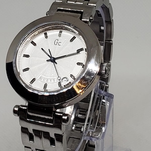 May include: A silver Guess watch with a white face and a silver band. The watch face has a circular pattern and the word "Guess" is printed on the face.