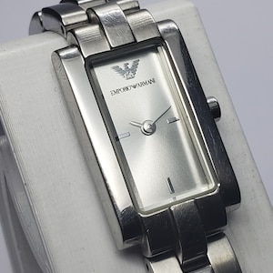 May include: A silver-toned Emporio Armani wristwatch with a rectangular face and a matching metal band. The watch face is silver with black hands and the Emporio Armani logo. The watch is displayed on a white surface.