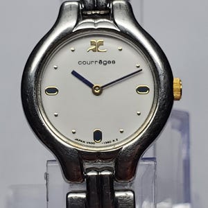 Puede incluir: Un reloj de pulsera Courr&egrave;ges plateado con esfera blanca, agujas negras y detalles dorados. El reloj presenta el logotipo de Courr&egrave;ges y el texto "JAPAN V400 -1360 R 2". La correa es plateada.