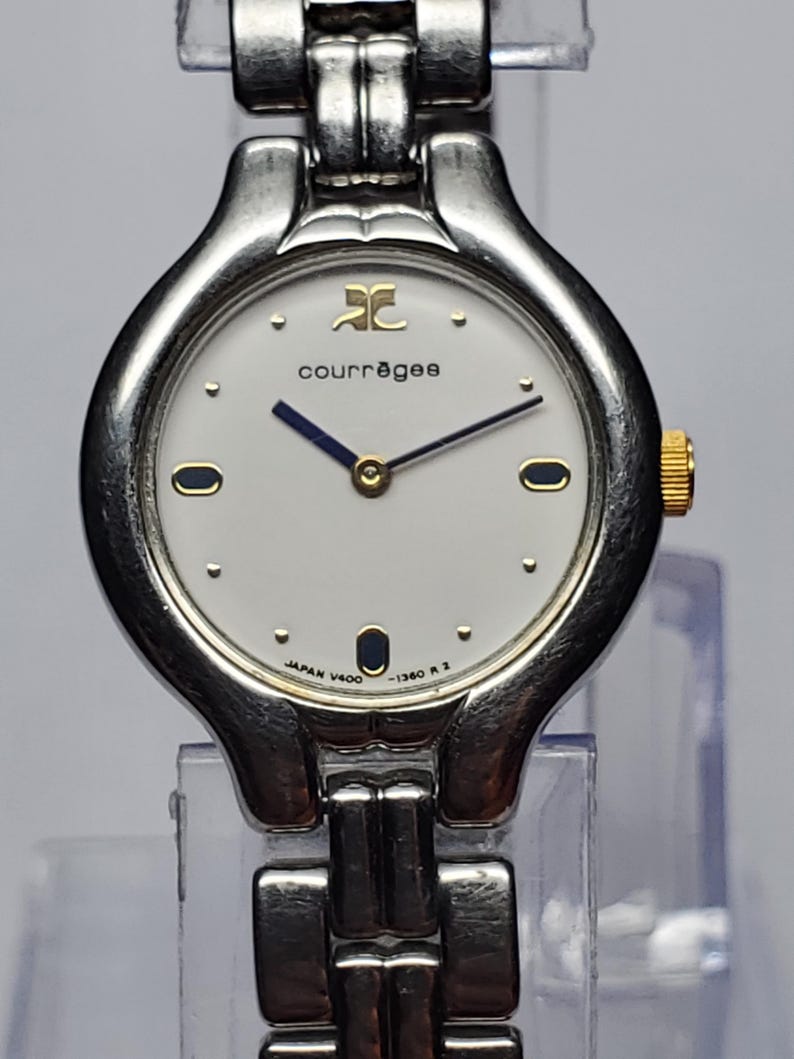 Puede incluir: Un reloj de pulsera Courr&egrave;ges plateado con esfera blanca, agujas negras y detalles dorados. El reloj presenta el logotipo de Courr&egrave;ges y el texto "JAPAN V400 -1360 R 2". La correa es plateada.
