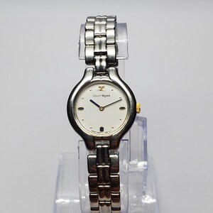 Puede incluir: Un reloj de pulsera Courr&egrave;ges plateado con esfera blanca y correa plateada. El reloj tiene una esfera redonda con detalles dorados y una pulsera de eslabones plateados. El reloj se exhibe sobre un soporte de pl&aacute;stico transparente.
