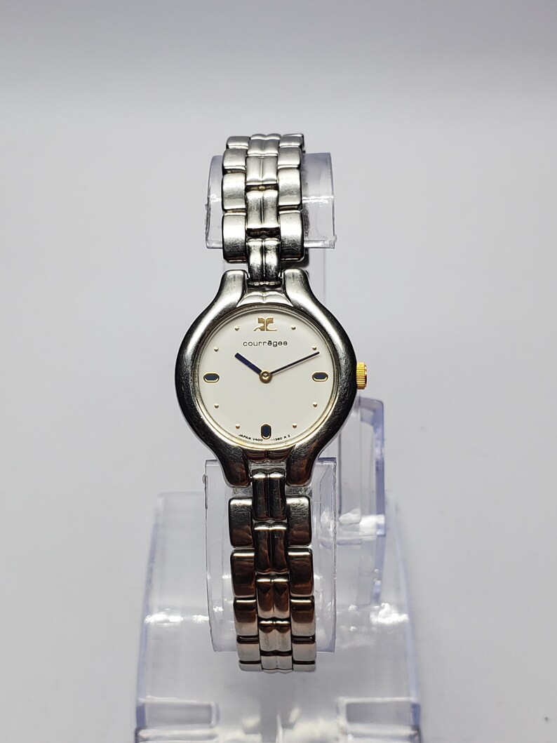 Puede incluir: Un reloj de pulsera Courr&egrave;ges plateado con esfera blanca y correa plateada. El reloj tiene una esfera redonda con detalles dorados y una pulsera de eslabones plateados. El reloj se exhibe sobre un soporte de pl&aacute;stico transparente.