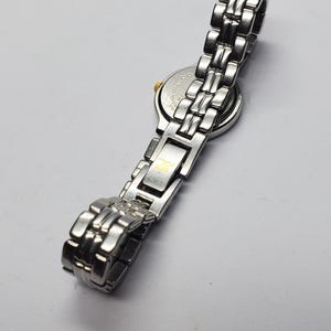 Puede incluir: Reloj de pulsera plateado con una correa de metal con eslabones. La esfera del reloj es redonda con un bisel dorado. La correa est&aacute; hecha de eslabones rectangulares. El reloj se muestra sobre una superficie blanca.