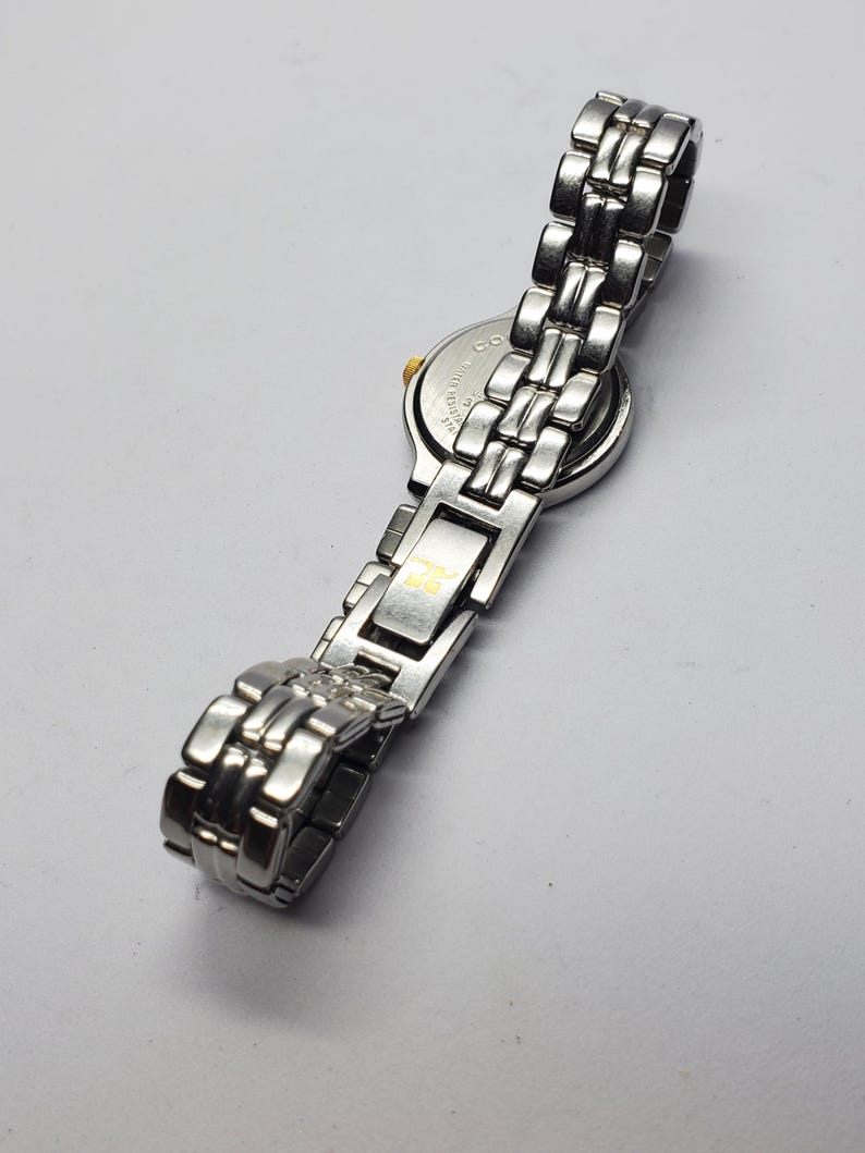 Puede incluir: Reloj de pulsera plateado con una correa de metal con eslabones. La esfera del reloj es redonda con un bisel dorado. La correa est&aacute; hecha de eslabones rectangulares. El reloj se muestra sobre una superficie blanca.