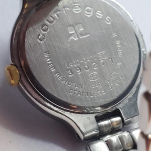 Puede incluir: Primer plano de un reloj Courr&egrave;ges de acero inoxidable. La parte trasera del reloj muestra el nombre de la marca, el logotipo y el texto que indica resistencia al agua y acero inoxidable. La correa del reloj es plateada.