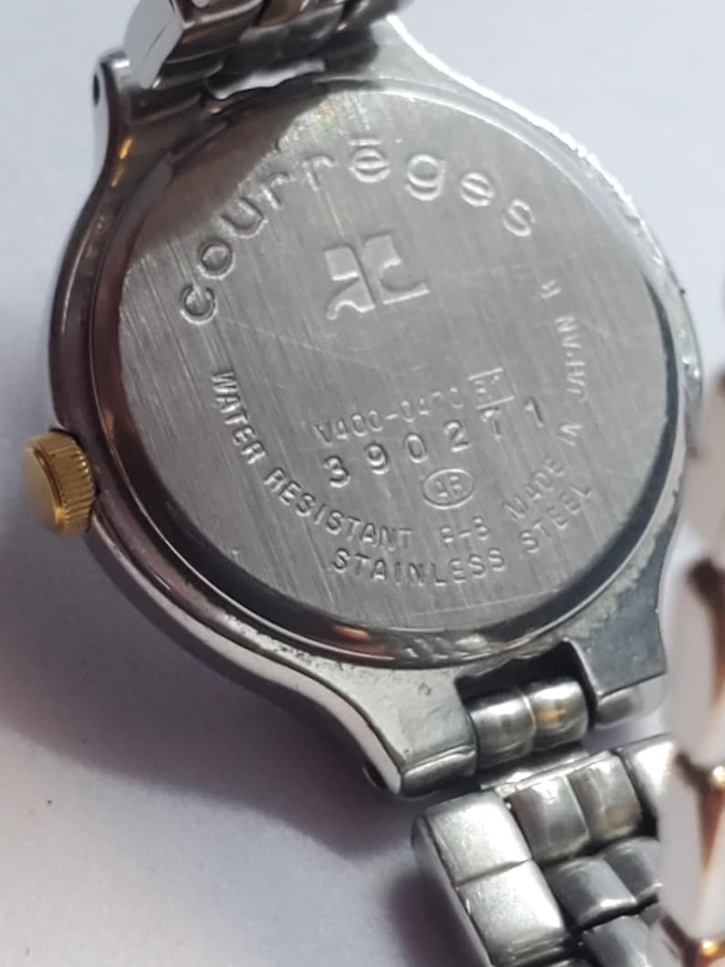 Puede incluir: Primer plano de un reloj Courr&egrave;ges de acero inoxidable. La parte trasera del reloj muestra el nombre de la marca, el logotipo y el texto que indica resistencia al agua y acero inoxidable. La correa del reloj es plateada.