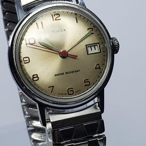 Könnte beinhalten: Eine silberfarbene Timex Armbanduhr mit champagnerfarbenem Zifferblatt, schwarzen arabischen Ziffern, einem Datumsfenster auf der Drei-Uhr-Position und einem flexiblen Metall-Zugband. Das Zifferblatt zeigt den Text Timex und Water Resistant.