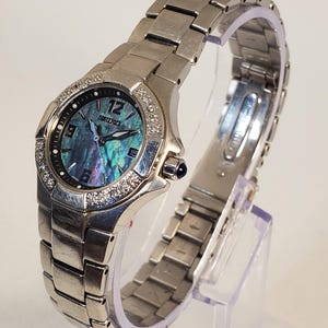 Puede incluir: Un reloj Seiko plateado con esfera de nácar y bisel con diamantes. El reloj tiene una correa de acero inoxidable y una indicación de fecha a las 3 en punto. El reloj se exhibe sobre un soporte transparente.