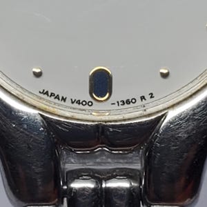 Puede incluir: Primer plano de un reloj de pulsera plateado con esfera gris claro. La esfera del reloj tiene un peque&ntilde;o &oacute;valo azul oscuro y el texto "JAPAN V400 -1360 R 2". La correa del reloj es plateada y parece ser de metal.