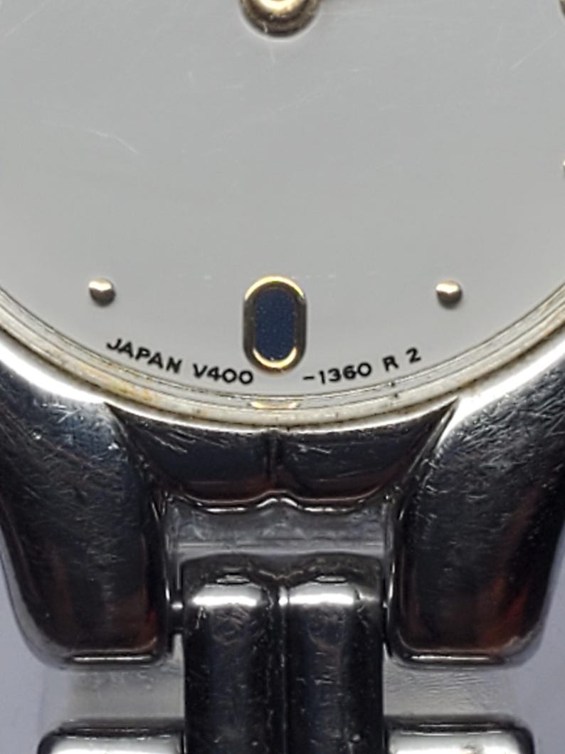 Puede incluir: Primer plano de un reloj de pulsera plateado con esfera gris claro. La esfera del reloj tiene un peque&ntilde;o &oacute;valo azul oscuro y el texto "JAPAN V400 -1360 R 2". La correa del reloj es plateada y parece ser de metal.