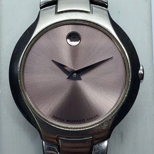 Pode incluir: Um relógio Movado em aço inoxidável com mostrador rosa e ponteiros pretos. O relógio apresenta um design minimalista com um único ponto às 12 horas e o texto "SWISS MOVADO MADE" ao redor do mostrador.