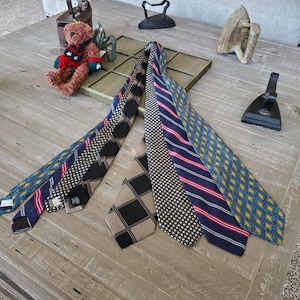 May include: A collection of six neckties with various patterns, including stripes, polka dots, and geometric designs. The ties are in shades of blue, black, and tan. The ties are displayed on a wooden table.