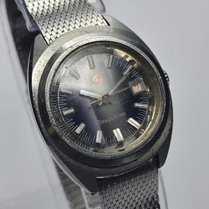 Op de afbeelding: Vintage SpaceWing polshorloge met een zilverkleurige mesh band en een donkerblauwe wijzerplaat. Het horloge heeft een ronde, zilverkleurige kast en een datumweergave. De wijzerplaat heeft de tekst "SPACEWING".