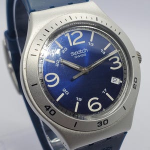 May include: A Swatch watch with a blue dial and a silver-tone bezel. The watch features large white numerals and hands, with a blue rubber strap. The watch face displays "SWISS".