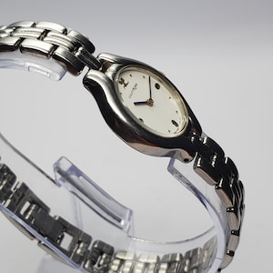 Puede incluir: Un reloj de pulsera Courr&egrave;ges plateado con una esfera ovalada blanca y agujas azules. El reloj presenta una correa de eslabones plateada y un dise&ntilde;o minimalista. El reloj se exhibe sobre un soporte de pl&aacute;stico transparente.