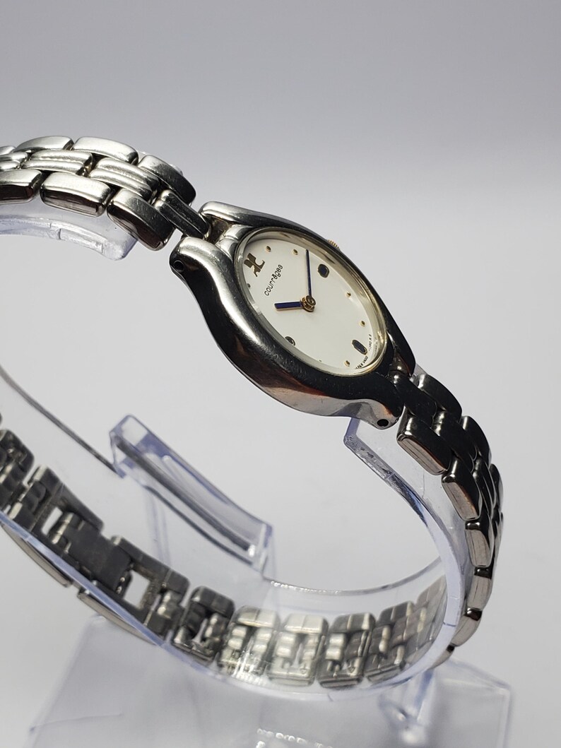 Puede incluir: Un reloj de pulsera Courr&egrave;ges plateado con una esfera ovalada blanca y agujas azules. El reloj presenta una correa de eslabones plateada y un dise&ntilde;o minimalista. El reloj se exhibe sobre un soporte de pl&aacute;stico transparente.
